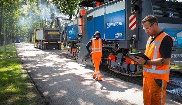 Roadwork scene with two workers in orange safety vests monitoring a large blue Wirtgen asphalt paving machine laying asphalt on a tree-lined street.
