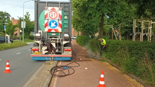 Road maintenance scene: a truck with traffic signs on its back, hoses on the ground, and a worker in a hi-vis vest trimming or spraying along the sidewalk by cones.