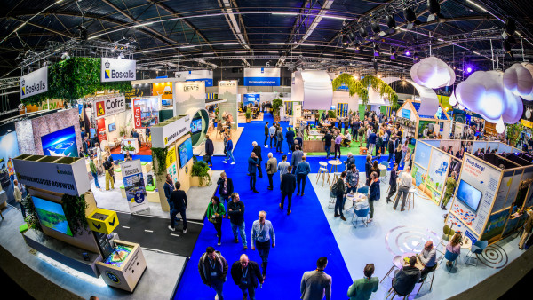 Wide shot of a crowded expo hall with blue carpet, rows of booths, banners, and attendees exploring displays.