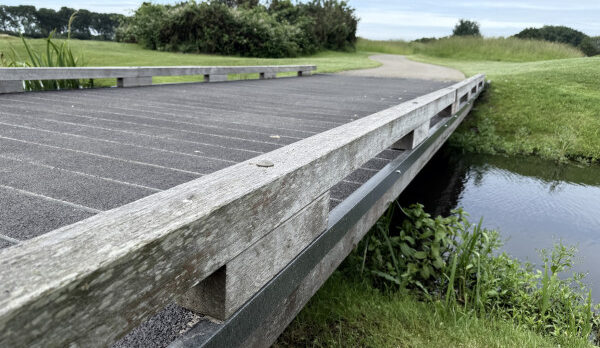 Wooden pedestrian bridge over a narrow canal in a grassy park, with a winding path in the background.